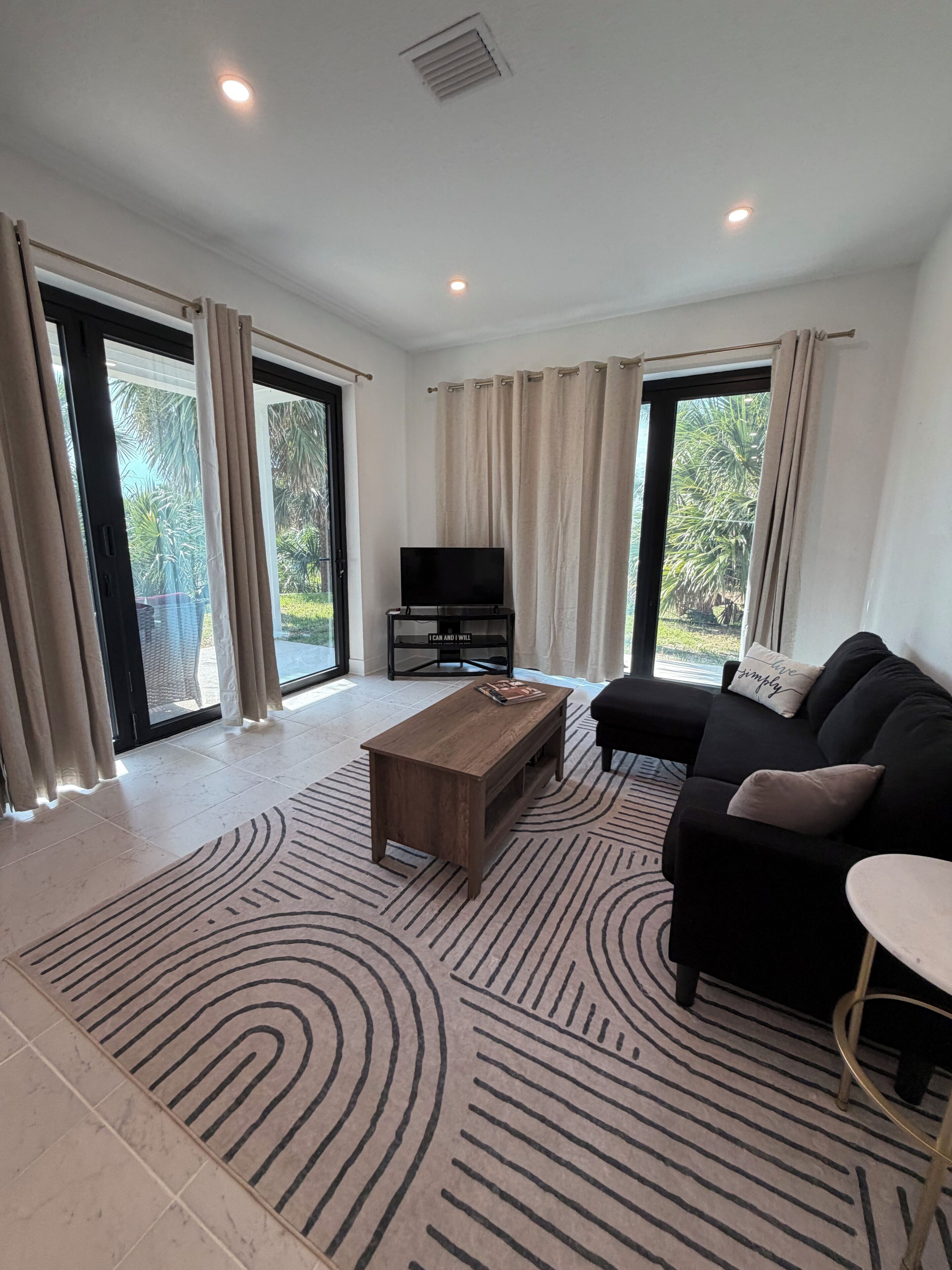 A spacious living room with a flat-screen TV, a wooden coffee table, a black sofa, and a patterned rug.