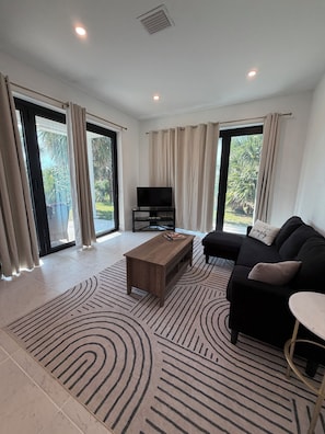 A spacious living room with a flat-screen TV, a wooden coffee table, a black sofa, and a patterned rug.
