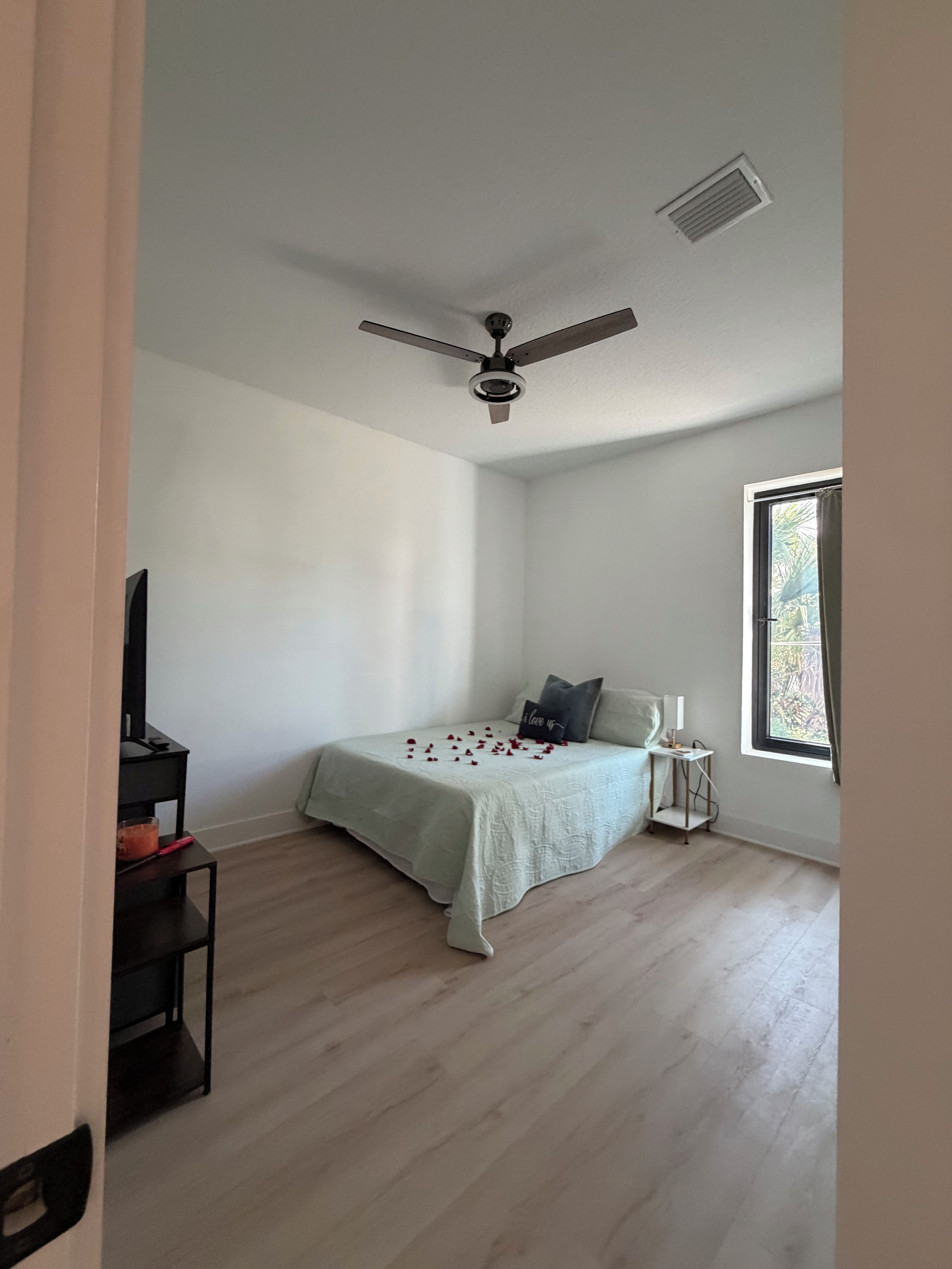 A bedroom with a bed, a ceiling fan, a window, and a small table with a lamp.