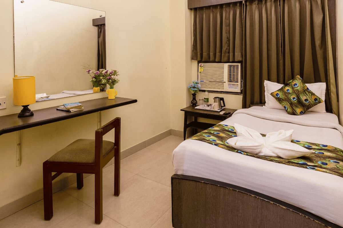 A hotel room with a bed, a desk with a chair, a mirror, a ceiling fan, and a window with curtains.