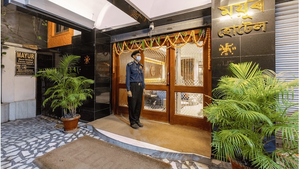 A hotel entrance with a security guard, a signboard with text, and potted plants. - Mayur Hotel (Guwahati)