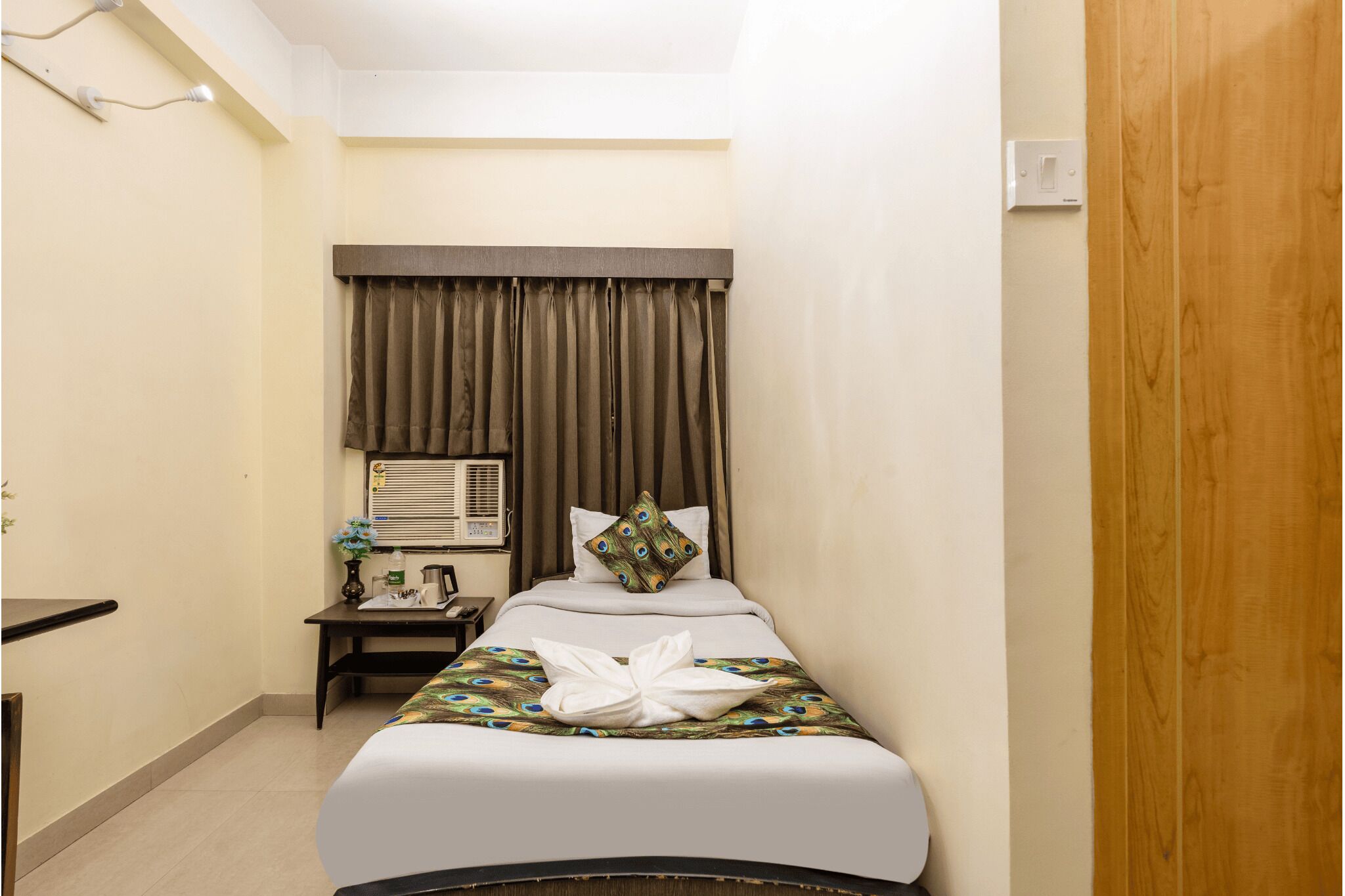A hotel room with a bed, a bedside table, an air conditioning unit, and a wooden door.