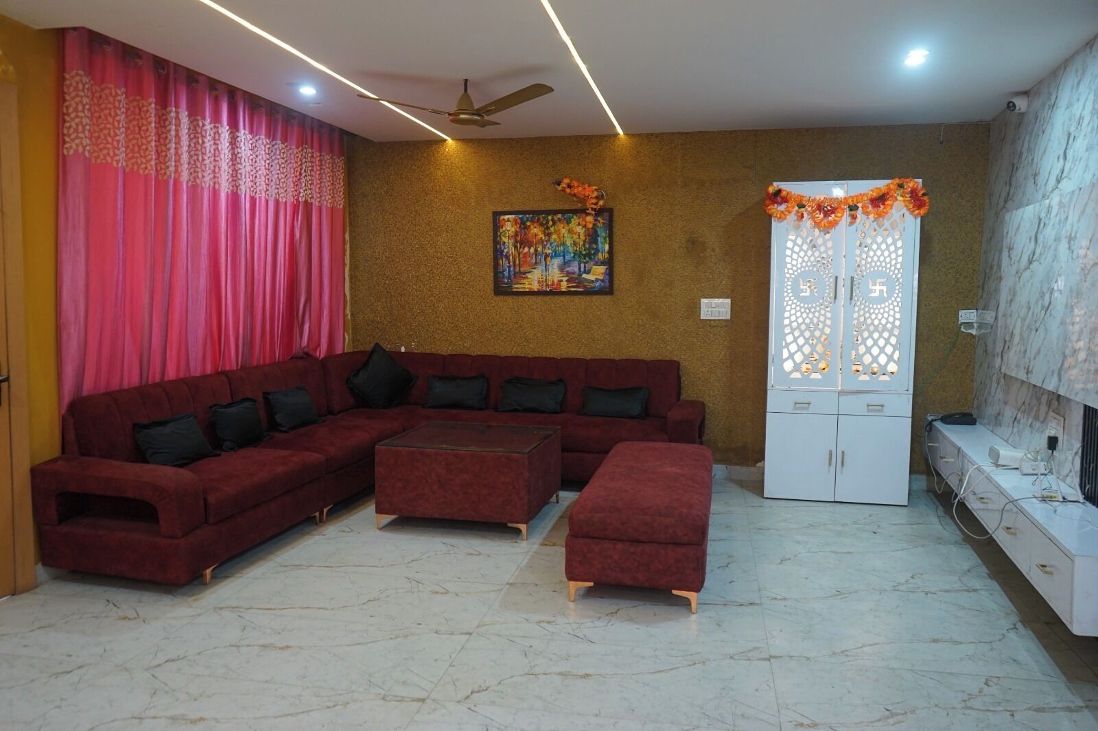 A living room with a sectional sofa, a coffee table, and an ottoman. There is a white cabinet with decorative elements on the wall, and a ceiling fan.