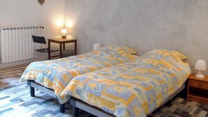 Ein Doppelbett mit Blumenmuster auf der Tagesdecke, ein Sessel, ein kleiner Tisch, eine Lampe und ein Heizkörper.