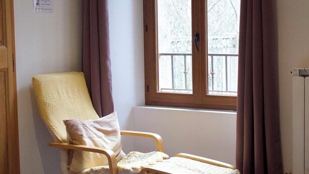 Ein Zimmer mit einem hölzernen Stuhl, einem Bett, einem Fenster mit Vorhängen und einem Heizkörper.