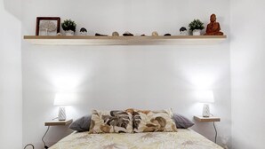 Una cama con ropa de cama floral, dos lámparas de noche, un estante con objetos decorativos y una pequeña estatua de Buda.