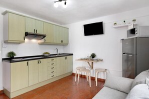 Cocina compacta con armarios verde claro, un refrigerador de acero inoxidable, un televisor de pantalla plana y una pequeña zona de comedor con taburetes.