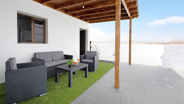 Un área de estar al aire libre con techo, con un sofá y sillas, una mesita con una planta y fruta, y vistas al paisaje.