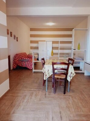 A dining area with a wooden table and chairs, a sofa with a checkered blanket, and a white shelving unit with bottles. - Residence Marino (Balestrate)