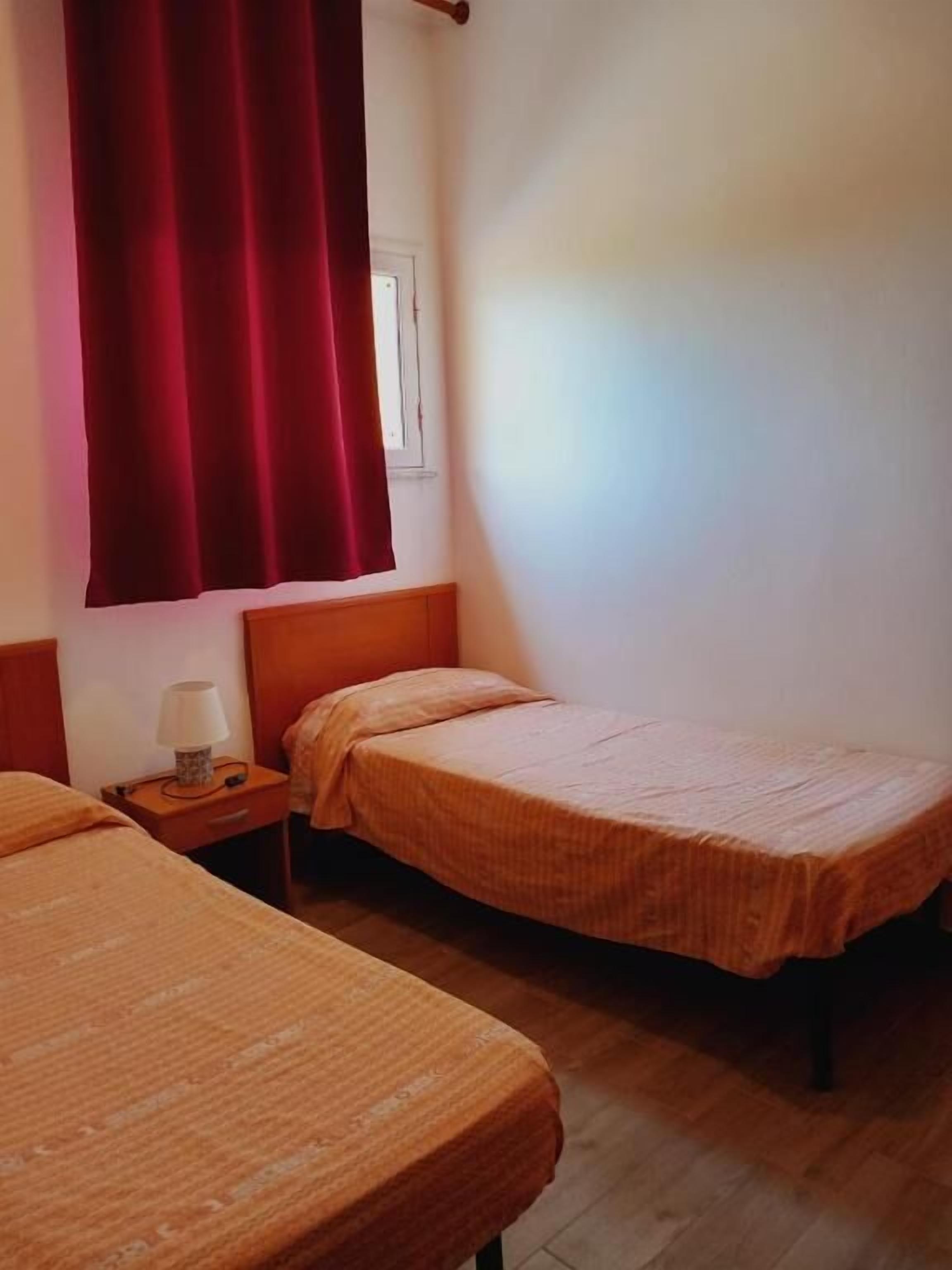 A hotel room with two beds, wooden headboards, a nightstand with a lamp, and a window with a red curtain.