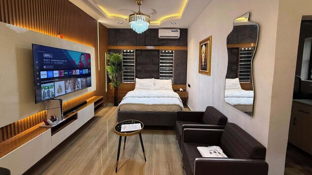 A modern hotel room with a large flat-screen TV, a bed, a sofa, a small table, and a mirror.