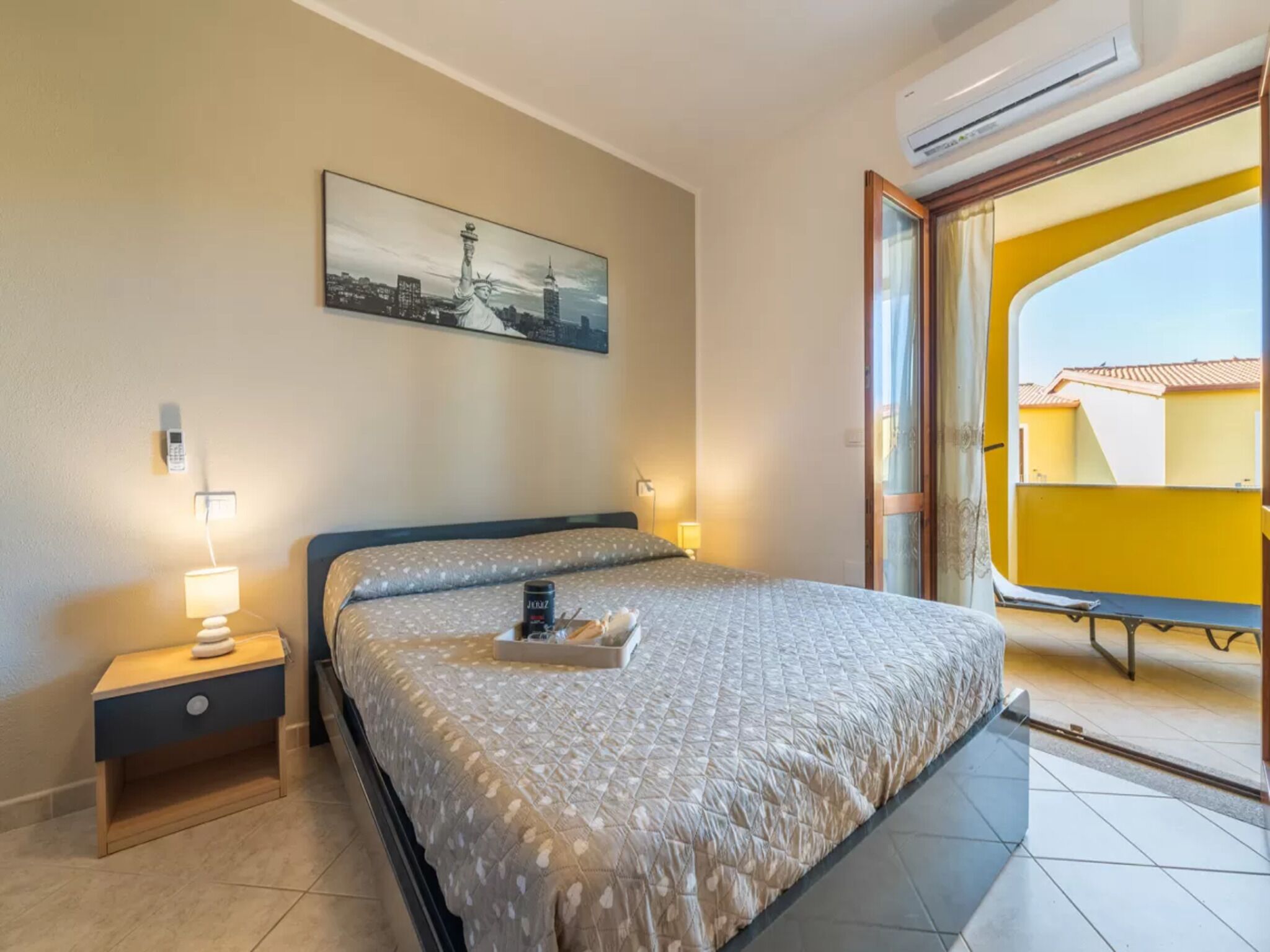 A bedroom with a bed, bedside lamps, a painting of the Statue of Liberty, and a balcony with yellow buildings.
