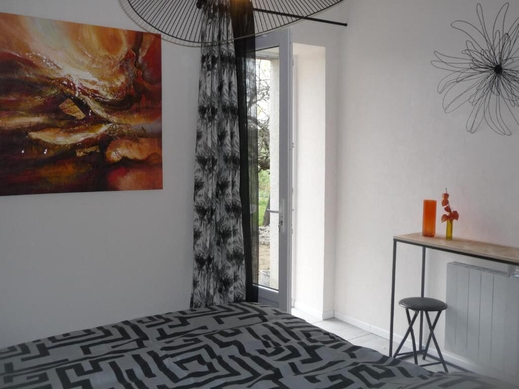 A modern bedroom with a geometric patterned bedspread, a large abstract painting, a black metal desk with a stool, and a view through a glass door.