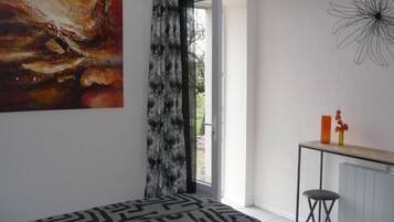 A modern bedroom with a geometric patterned bedspread, a large abstract painting, a black metal desk with a stool, and a view through a glass door.
