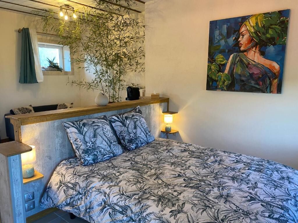 Un dormitorio con cama, mesita de noche, lámpara, ventana con cortinas, un cuadro en la pared y una planta.
