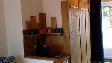 A room with wooden cabinets, a small shelf with cups, a potted plant, and a window with a curtain.