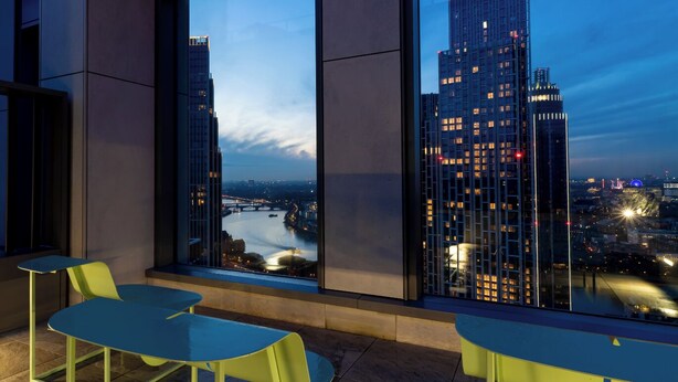 Un interior moderno con un ventanal grande que da a una vista panorámica de la ciudad al atardecer, con dos mesas y un banco.