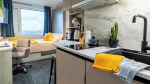 Una habitación de hotel moderna con kitchenette, una cama con almohadas amarillas, un escritorio con silla, una ventana con cortinas y vista a la ciudad.