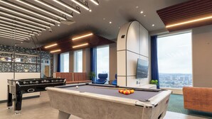 Una mesa de billar con una mesa de fútbol de mesa y un televisor en una habitación moderna con ventanas amplias y vista a la ciudad.
