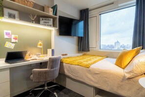 Une chambre à coucher comprenant un lit, un bureau avec un ordinateur portable, une chaise, un téléviseur et une fenêtre donnant sur la ville.
