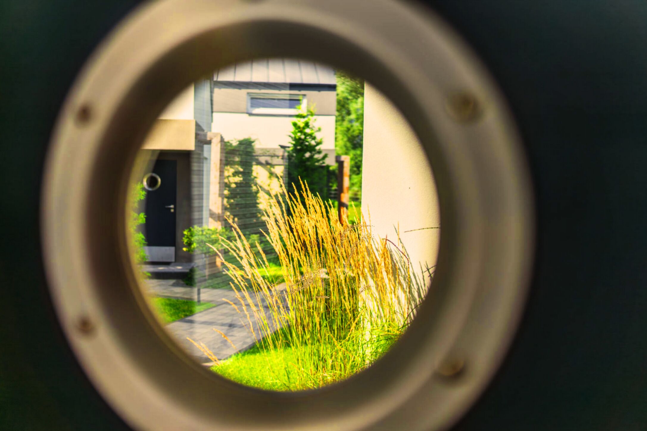 A view through a circular opening, revealing a building with a modern design, a door, and some grass in the foreground.