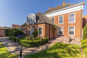 A two-story brick house with a sloped roof, a small garden, and a paved driveway. - "Cirksena" Vacation Apartment - Spacious living area with garden access (Krummhörn)