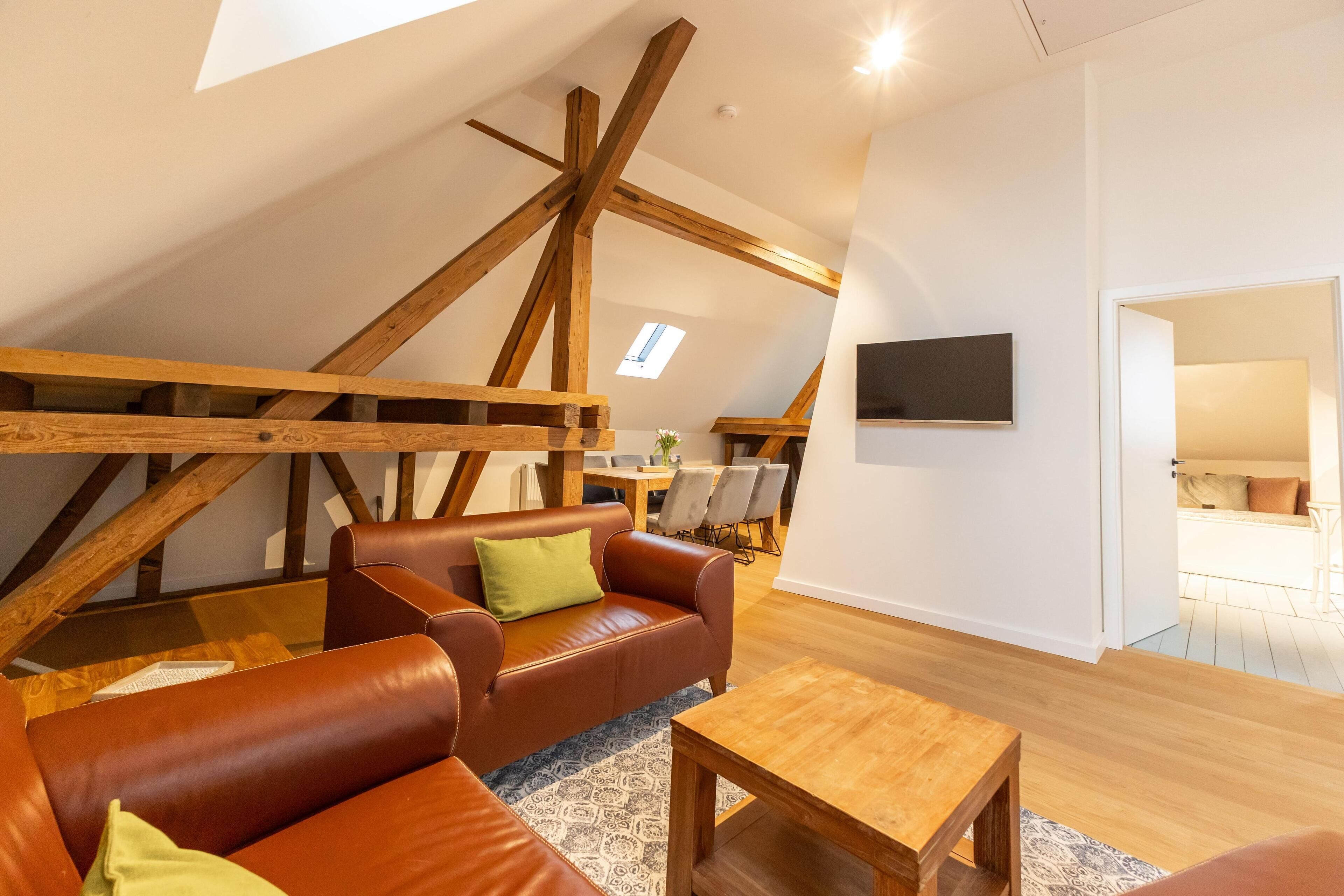 Ruime loft slaapkamer met zichtbare houten balken, een groot raam, een bruine leren bank, een houten salontafel en een flatscreen televisie aan de muur bevestigd.