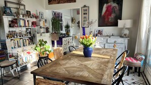 Zona pranzo con tavolo di legno, sedie in vimini, un vaso blu con fiori e una libreria piena di libri e oggetti decorativi.