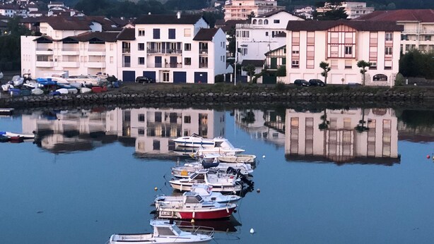 Un porto turistico con diverse imbarcazioni ormeggiate, circondato da edifici con balconi e uno sfondo montuoso.
