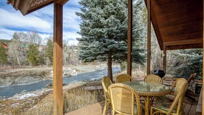 Une terrasse en bois, meublée de mobilier en osier, donnant sur une rivière et un arbre recouvert de neige.