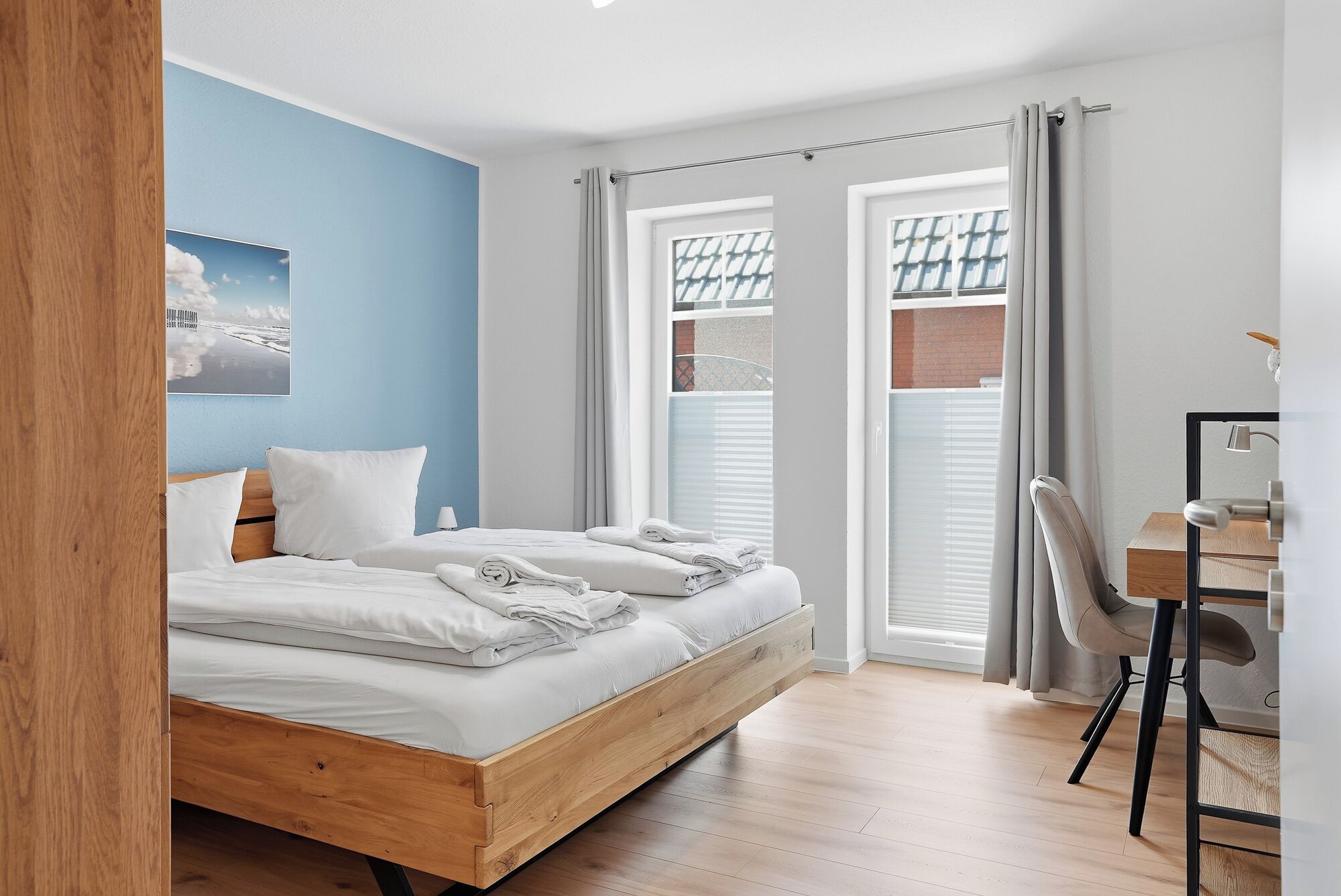 A bedroom with a wooden bed, a desk, and a view of the outside through a window with blinds.