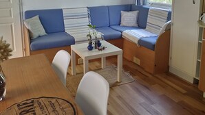 A cozy living area with a blue sofa, a small table with decorative items, a white cabinet, and a window with curtains. - Seasonal rental fully equipped mobile home for 4/6 people (Saint-Hilaire-de-Riez)