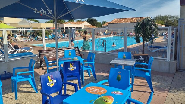 A poolside area with blue chairs and tables, a large umbrella, and a pool with people swimming and lounging. - Seasonal rental fully equipped mobile home for 4/6 people (Saint-Hilaire-de-Riez)