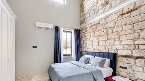 Un dormitori amb una paret de pedra, un llit amb capçalera blava, una panxa vermella i una finestra amb cortines.