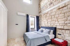 A bedroom with a stone wall, a bed with a blue headboard, a red bench, and a window with curtains.