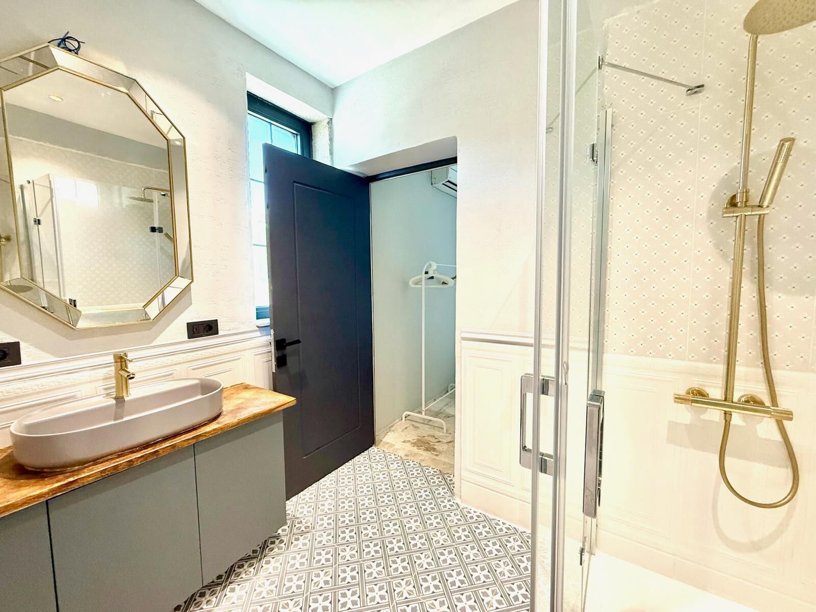 A bathroom with a large mirror, a sink with a gold faucet, a shower with a gold showerhead, and patterned floor tiles.