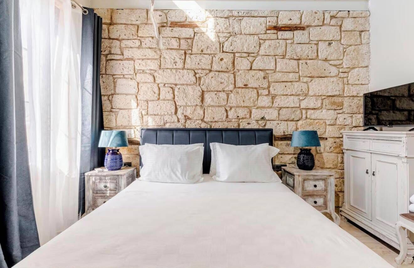 Un dormitorio con pared de piedra, una cama con ropa de cama blanca, dos mesitas de noche con lámparas y un armario.