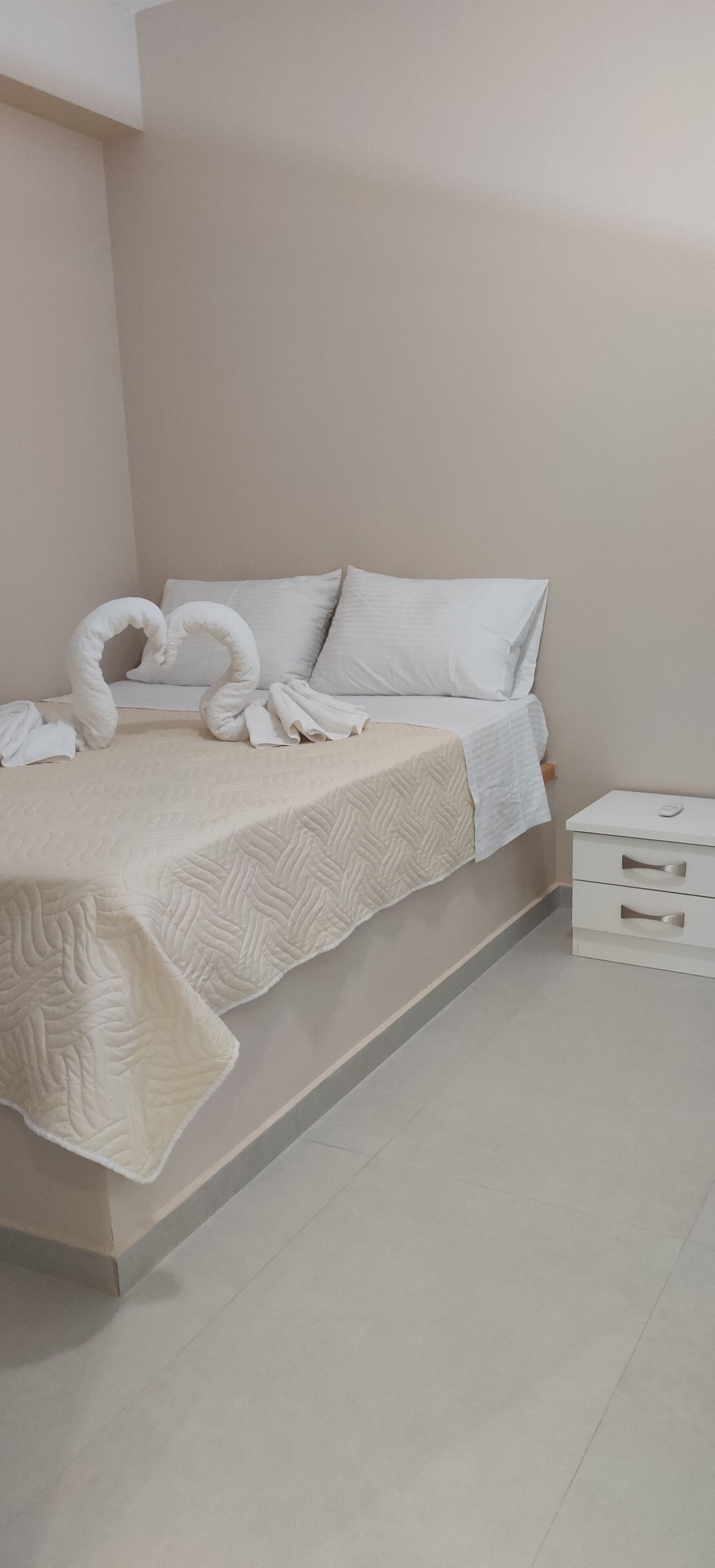 A neatly made bed with a quilted cover, a heart-shaped towel decoration, and a white bedside table with drawers.