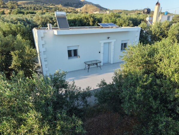 Un bâtiment blanc doté de panneaux solaires, une petite table et une porte, le tout entouré d’une végétation luxuriante.