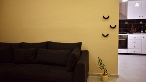 Un salon avec un canapé de couleur sombre, une petite plante en pot et une cuisine visible à travers une arche.