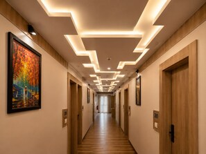 Un corridoio d'albergo con porte in legno, cartelli con i numeri delle camere e un soffitto con lampade geometriche.
