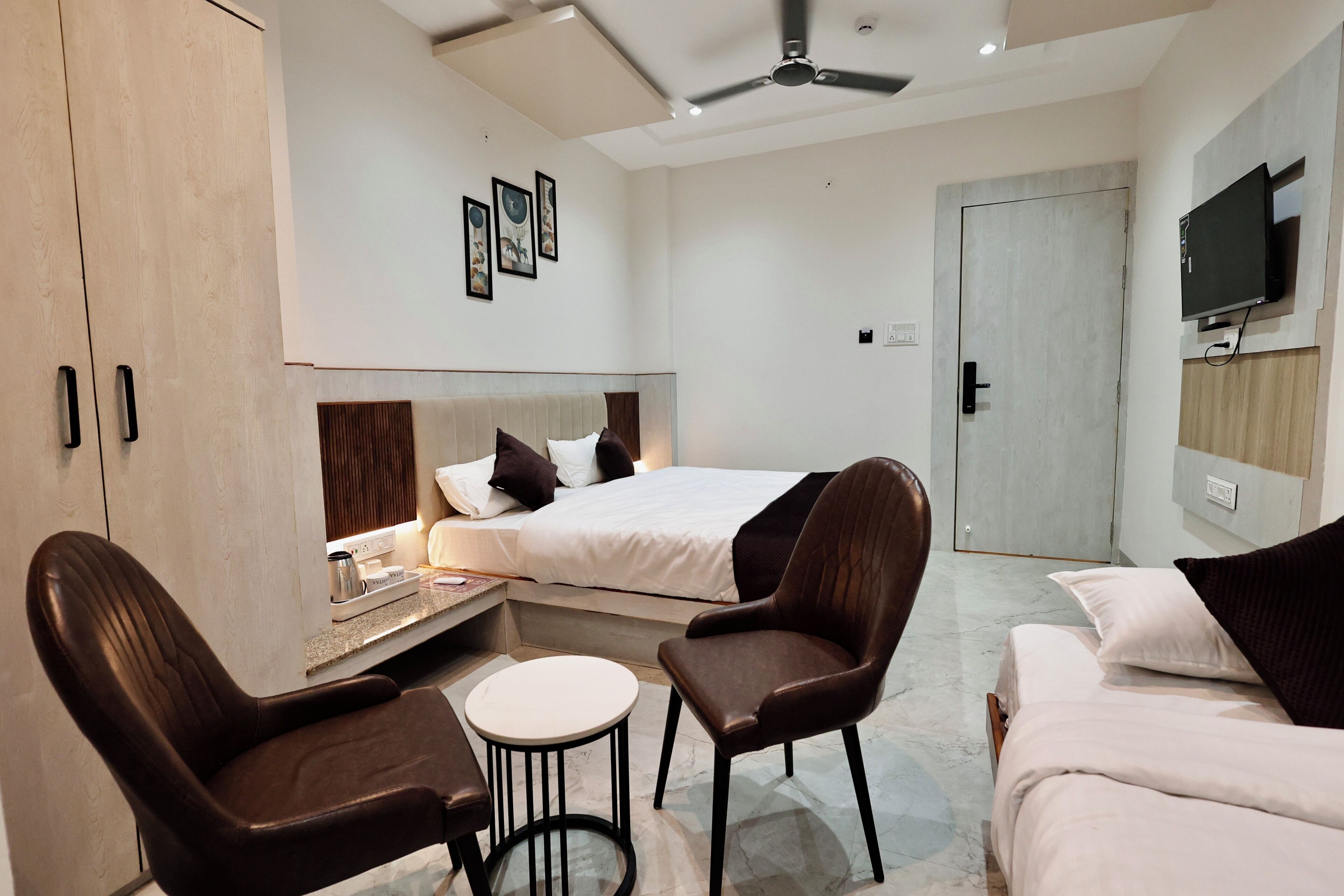 A modern hotel room with a bed, two chairs, a small table, a ceiling fan, and a TV mounted on the wall.