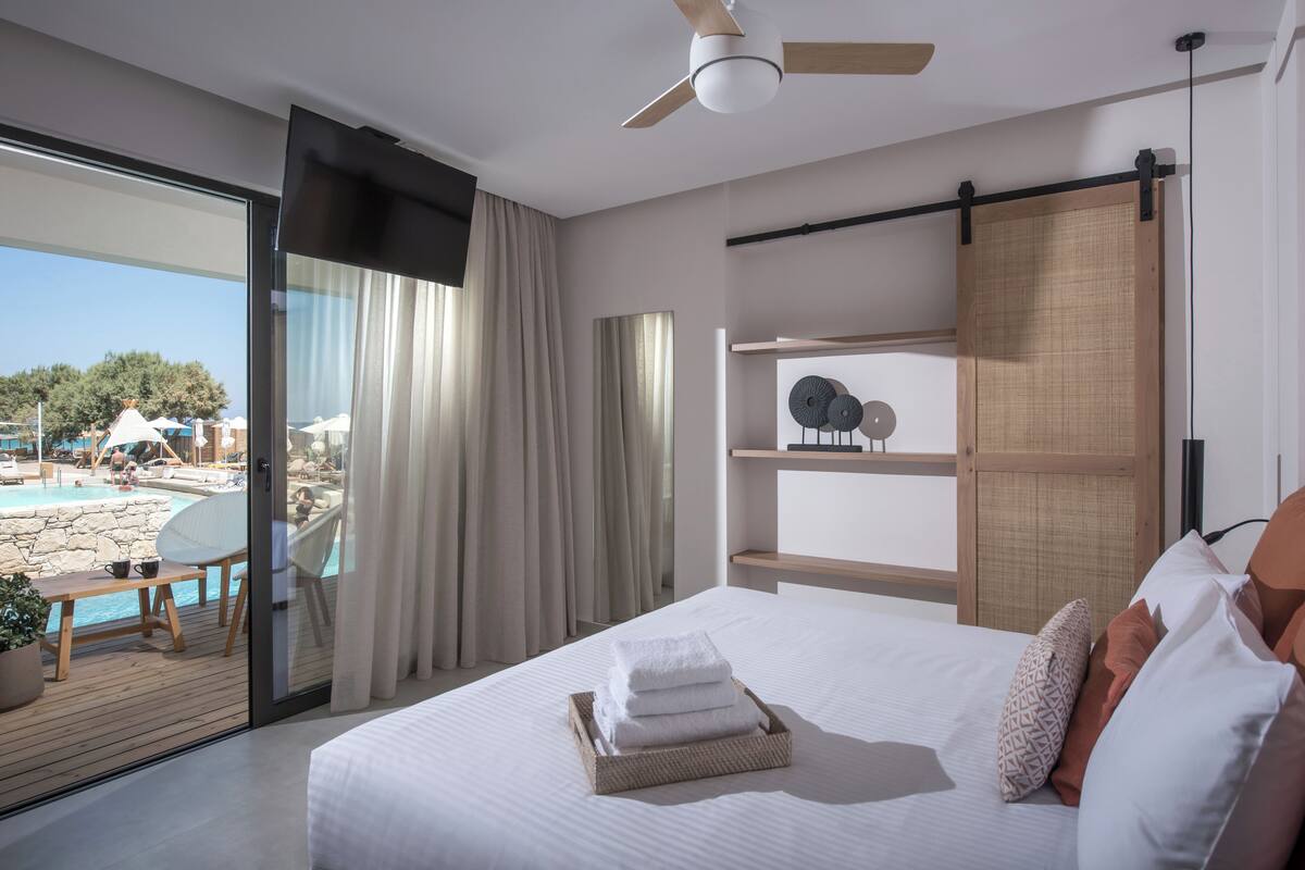 Ein modernes Hotelzimmer mit einem großen Bett, einem Deckenventilator, Blick auf Pool und Strand sowie einer Schiebetür zum Balkon.