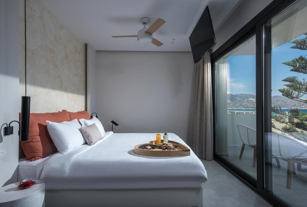 A modern hotel room with a large bed, a ceiling fan, a tray of breakfast items, and a balcony with a view of the city.