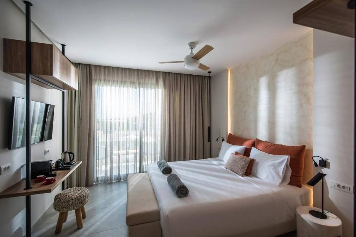 Ein modernes Hotelzimmer mit einem großen Bett, einem Schreibtisch mit Fernseher, einem Deckenventilator und einem großen Fenster mit Vorhängen.