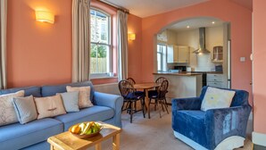 A living room with a sofa, a dining table, and chairs. There is a kitchen visible through an archway. - Captain's Cottage at Mixtow Creek (Lanteglos)