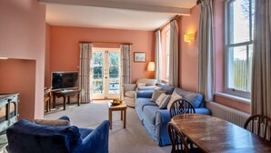 A living room with a sofa, a TV, a table with a fruit bowl, and a window with curtains. - Captain's Cottage at Mixtow Creek (Lanteglos)
