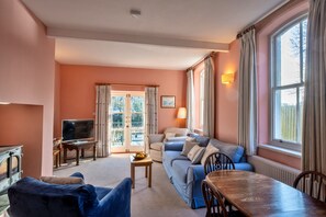 Ein Wohnzimmer mit einer Couch, einem Fernseher, einem Tisch mit Obstschale und einem Fenster mit Vorhängen.