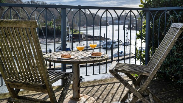 A wooden deck with two chairs and a table, overlooking a body of water with boats. - Captain's Cottage at Mixtow Creek (Lanteglos)