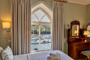 Ein Hotelzimmer mit einem großen Erkerfenster, einer Holzkommode mit Spiegel und einem Bett mit weißer Bettdecke.